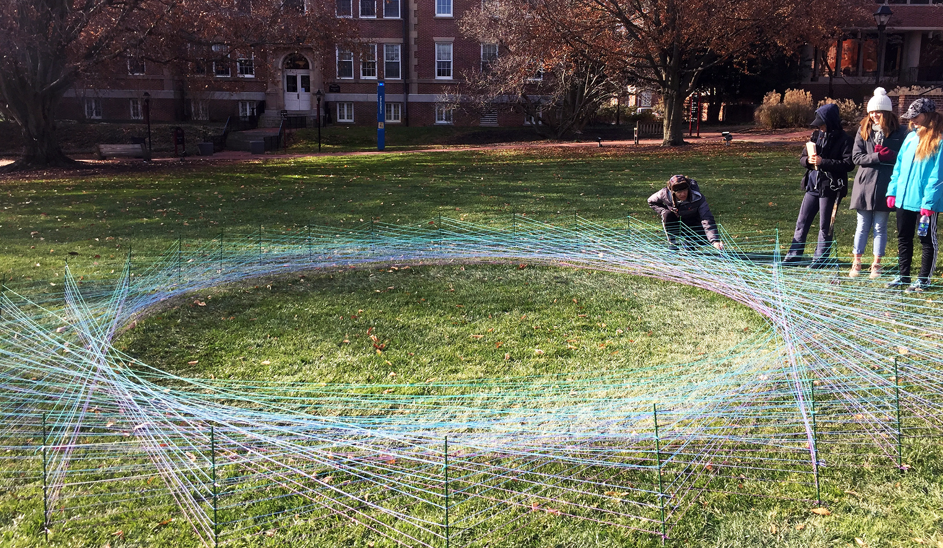 Students gathered around a student artwork made of colorful intersecting strings, in front of Bunting Hall on the WC campus