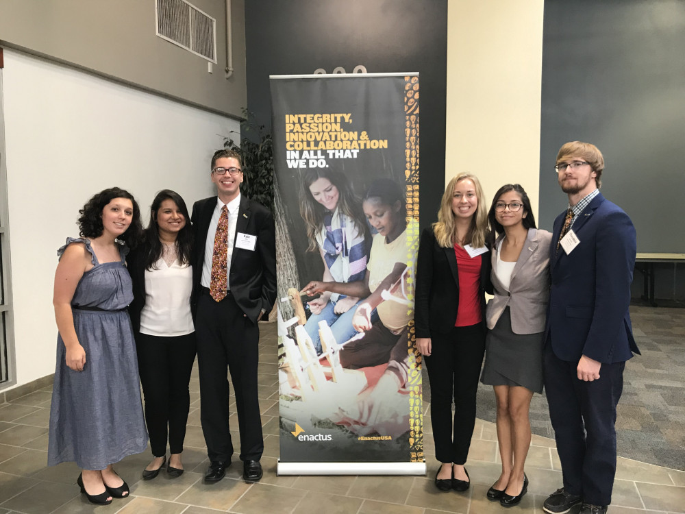 Four female and two male student members of Enactus stand in front of an Enactus banner. They are formally dressed.