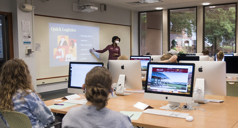 Students at computers look to an instructor at the front of the room going over a slide labeled "Quick Logistics"