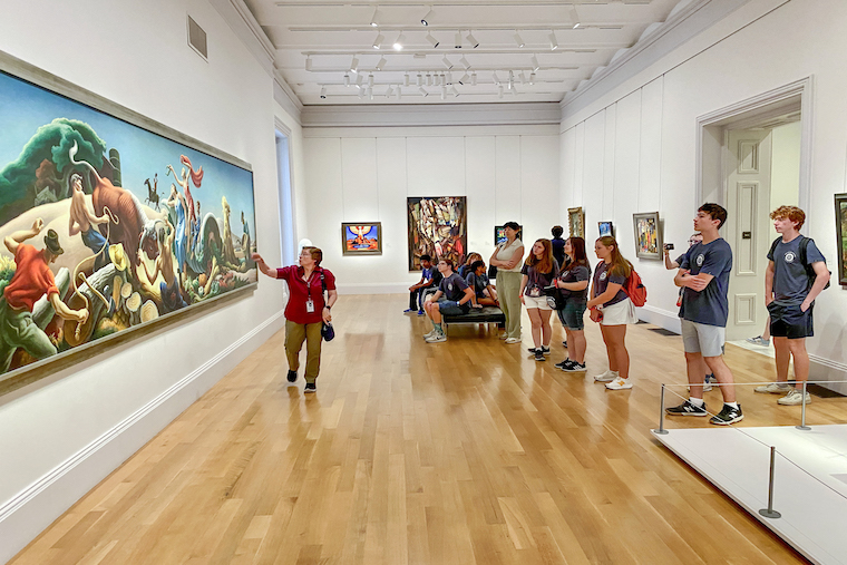 Students in an art gallery listen to an instructor gesturing toward a painting