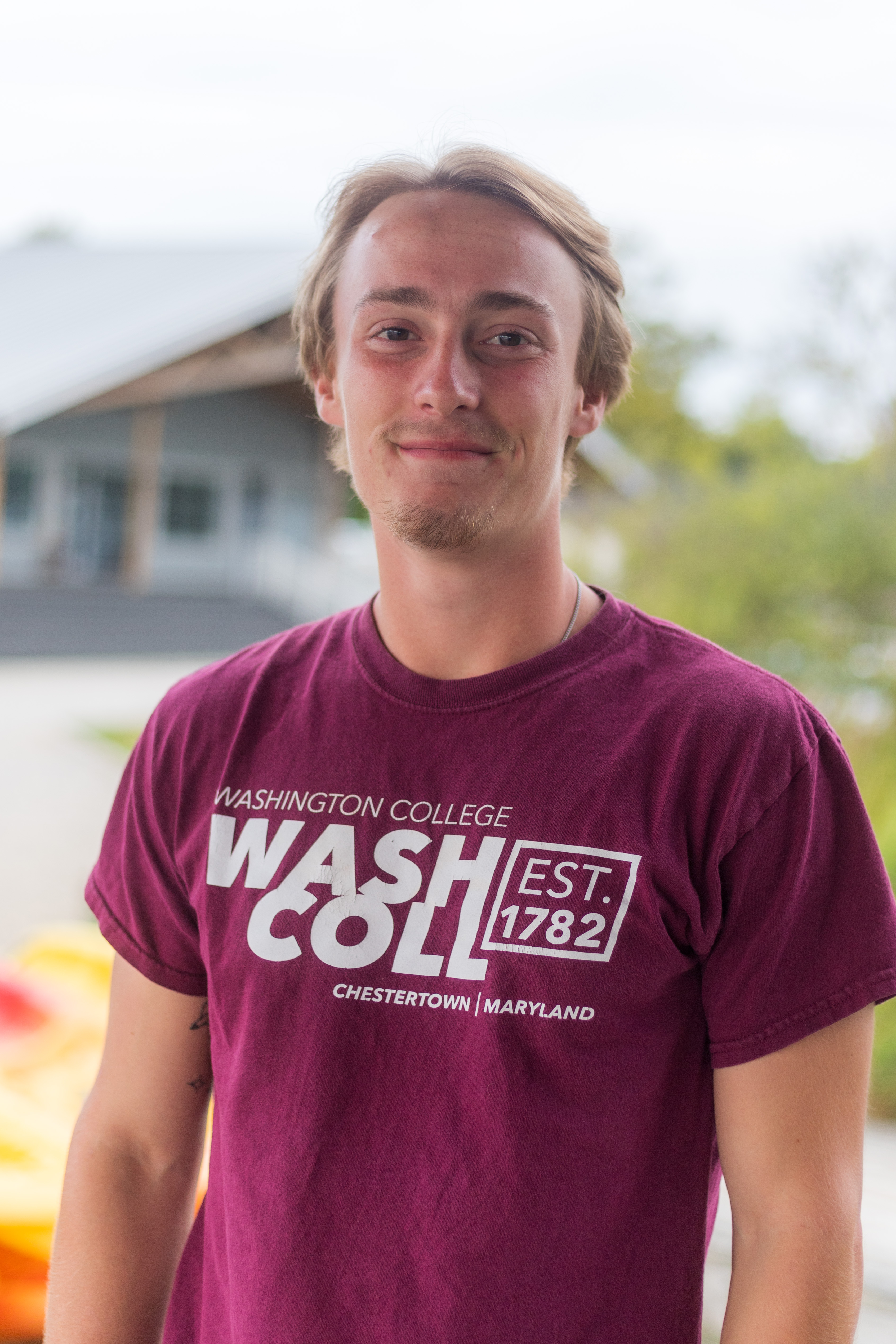 Adam, wearing a Washington College t-shirt and smiling, with the Boathouse in the background. 