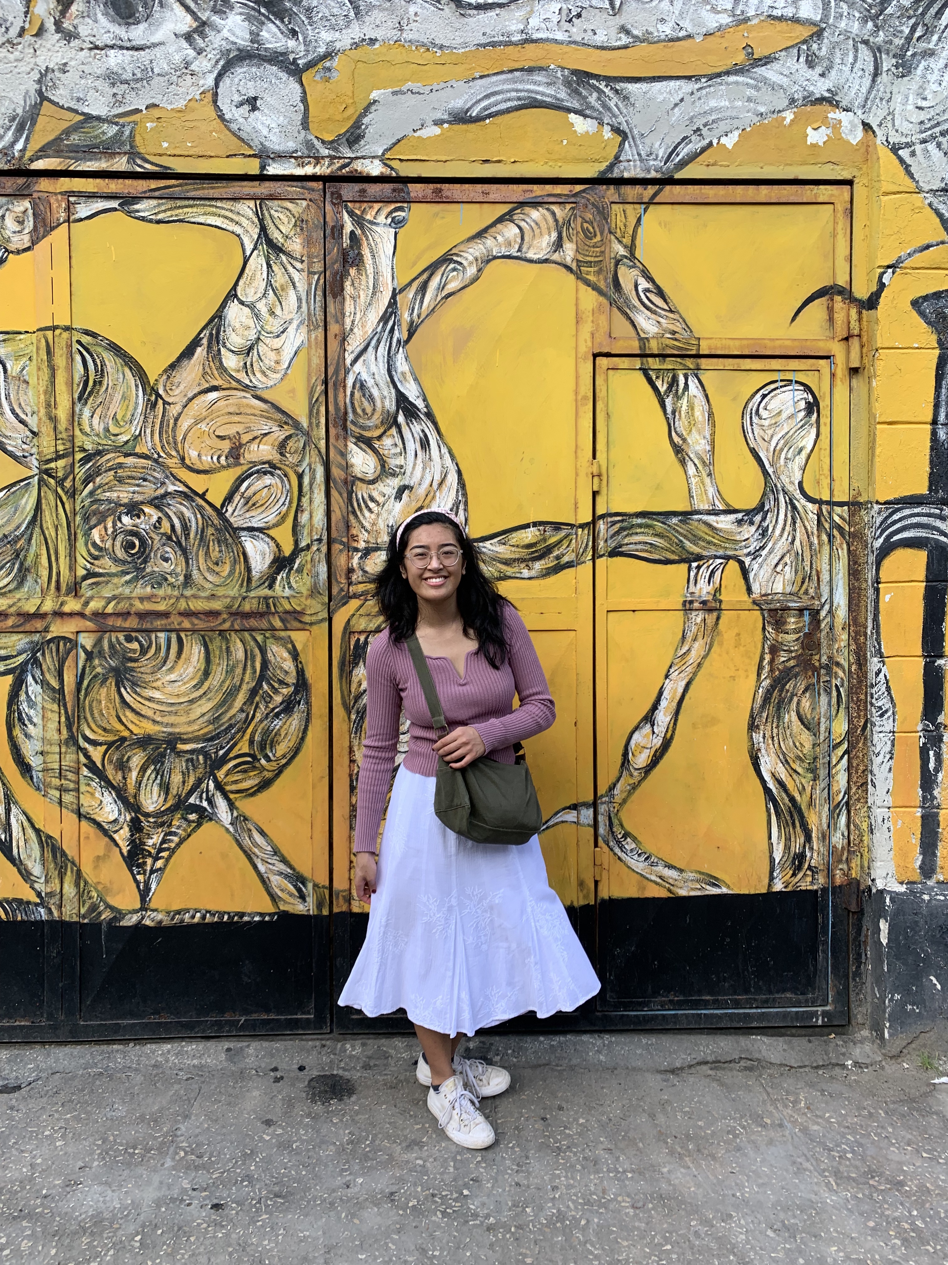 Regina, wearing a pink top and long white skirt, posing in front of a yellow and white mural depicting human figures in Havana, Cuba. 