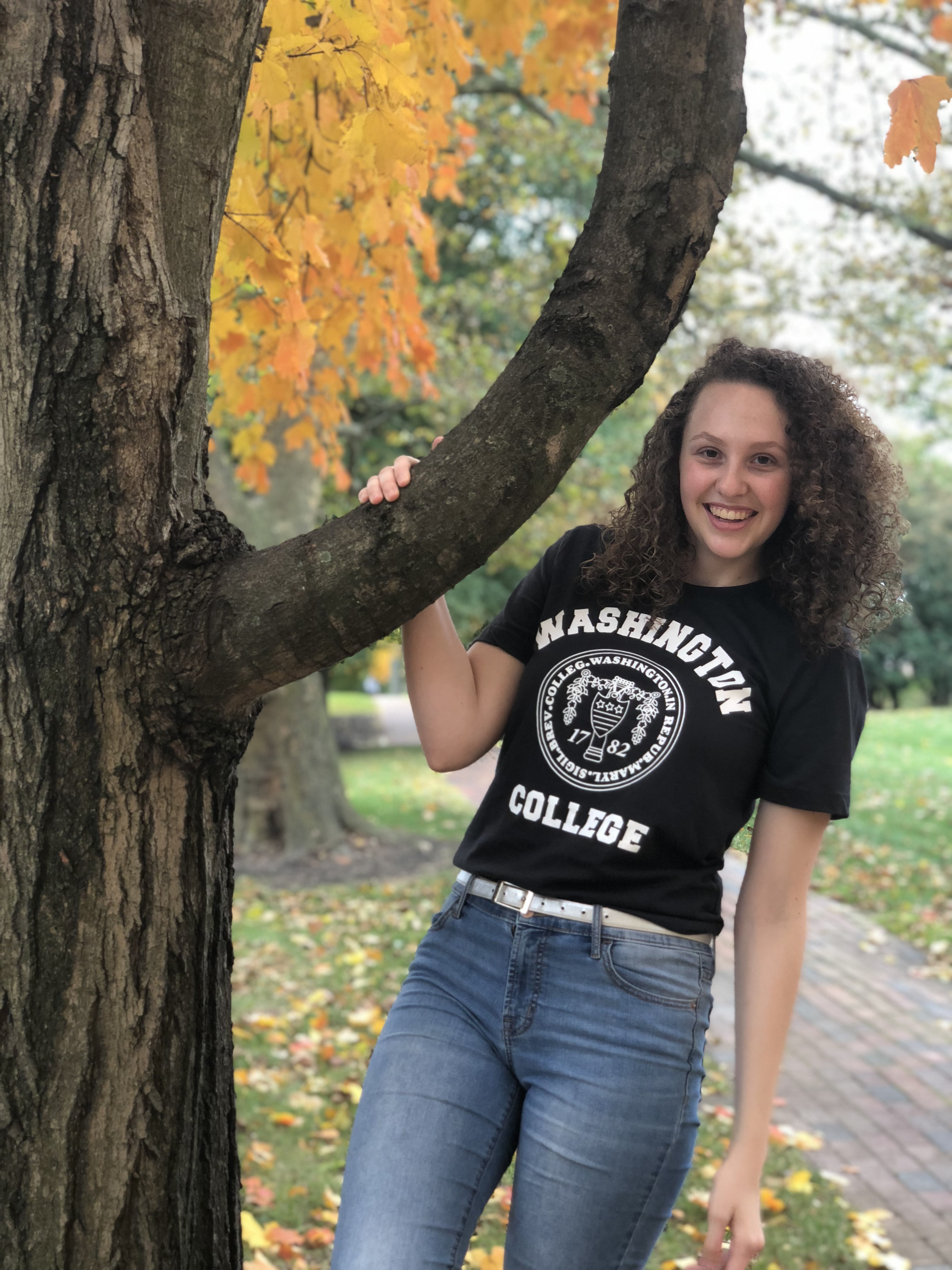 Carlee, smiling and wearing a black t-shirt with the Washington College logo, blue jeans, and a white belt in front of fall foliage. 