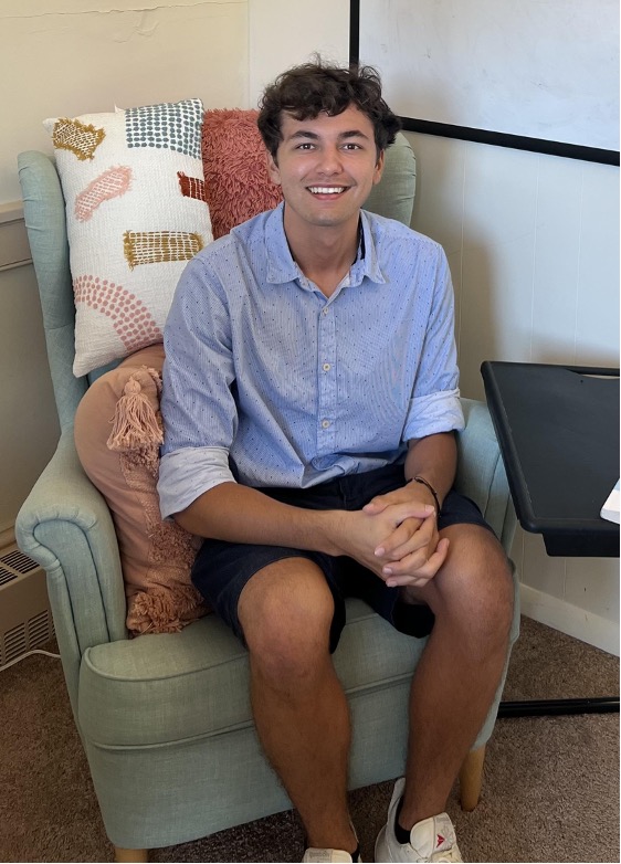 Noah, smiling and wearing a blue shirt and navy shorts, sitting on a blue chair with decorative pillows. 